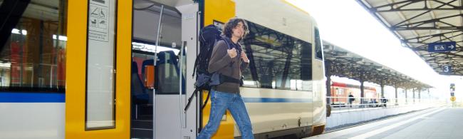 A covered track at a railway station. A man with a large backpack gets off a train with yellow doors.