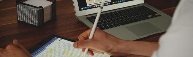 Arbeitsplatz mit Laptop, iPad, Notizzettelbox und Hand die einen Stift hält