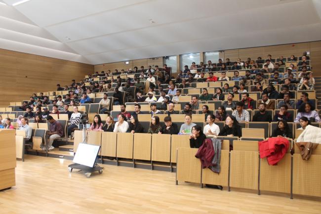 Ein mit etwa 150 sitzenden Personen gefüllter Hörsaal mit Sitzbänken aus hellem Holz