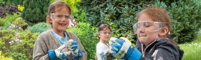 Kinder hocken auf einer Wiese, sie tragen Schutzbrillen und Arbeitshandschuhe