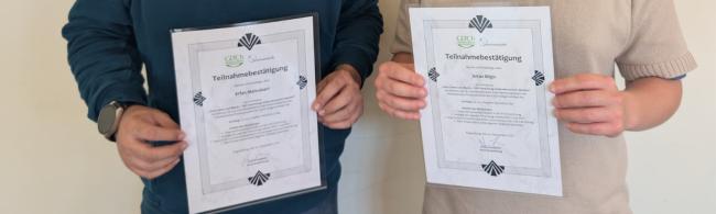 Zwei Doktoranden stehen nebeneinader und zeigen ihr Teilnahmezertifikat in die Kamera
