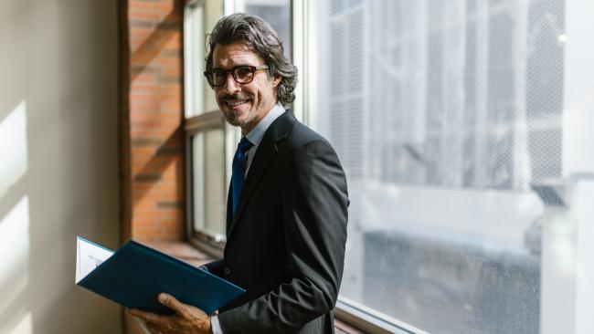 Ein Mann in schwarzem Anzug, der seitlich vor einer großen Fensterfront steht.