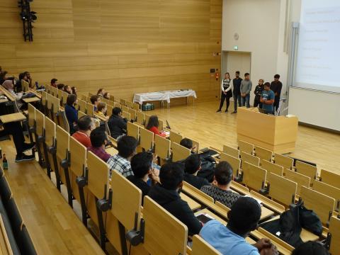 Blick aus dem Auditorium auf die Bühne eines Hörsaals, Studierende auf der Bühne und im Auditorium