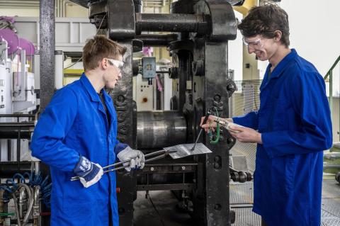Zwei Männer im Blaumann stehen vor einer Walze. Einer hält ein Blech der andere ein Messgerät