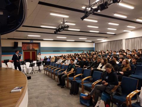 Ein Mann in einem großen Vortragssaal an einer Schule. Die Zuhörerschaft besteht aus jugendlichen Schülerinnen und Schülern.