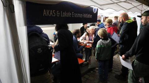 Beleuchtete Stände mit vielen Besuchern, Standbeschriftung: &quot;AKAS - Lokale Erasmus-Initiative&quot;
