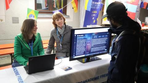 Zwei Frauen an einem Stand mit Computerbildschirm und Notebook führen einem Besucher etwas vor