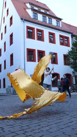 Ein Frau schwingt ein gold-gelbes Band an einer Leine auf einem Platz