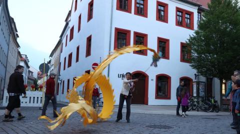 Ein Frau schwingt ein gold-gelbes Band an einer Leine auf einem Platz