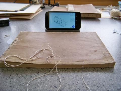 Eine Nahaufnahme eines Arbeitstisches in einer konservatorischen Werkstatt. Im Zentrum liegt ein altes, ungebundenes Buch mit braunem Umschlag. Ein heller Faden liegt bereit, um den Buchblock neu zu heften. Direkt dahinter liegt ein Smartphone, das ein schematisches Diagramm oder eine digitale Anleitung für den nächsten Arbeitsschritt anzeigt.