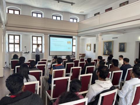 Ein Vortrag im Senatssaal der TUBAF, mehrere Studierende sitzen auf weiß lackierten Holzstühlen mit roten Polsterbezügen und verfolgen einen Vortrag (männlicher Referent) mit Präsentation (Videoprojektor auf Leinwand)