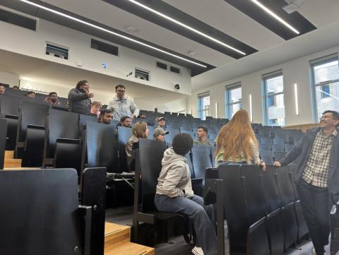 Studierende in einem Hörsaal mit starkem Gefälle, einige stehen und lachen, die meisten sitzen, die Sitzbänke sind schwarz und aus Holz, es herrscht gute Stimmung
