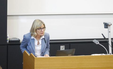 Eine blonde Frau mit Brille steht an einem Präsentationstisch in einem Hörsaal und spricht.