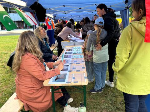 Ein Stand zur Nacht der Wissenschaft auf einer Wiese. Auf dem Tisch liegen laminierte Seiten. Besucher stehen davor, auf einer Bank sitzen Frauen, die den Stand betreuen.