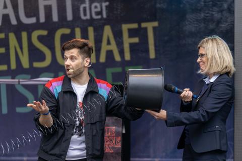 Ein Bühnenexperiment mit einem Moderator und eine Kandidatin, die ein Mikrofon in einen Eimer hält.
