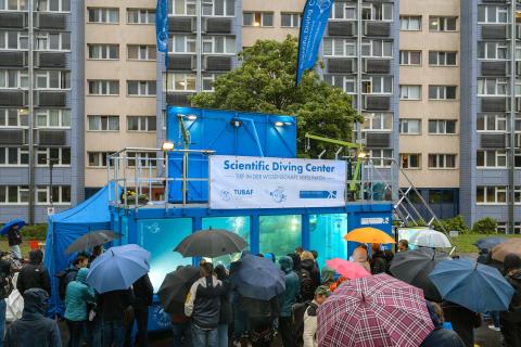 Ein Tauchcontainer, davor Menschen mit Regenschirmen