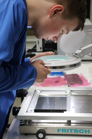 Ein Studierender kontrolliert die Qualität des Lötpastenauftrags auf einer PCB nach dem Schablonendruck im SMT-Fertigungsprozess.
