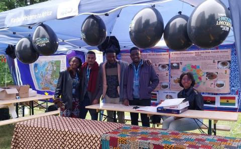Gruppenfoto mit fünf Studierende aus Afrika (Frauen und Männer) in einem Stand mit Plakaten zum Thema afrikanische Küche.