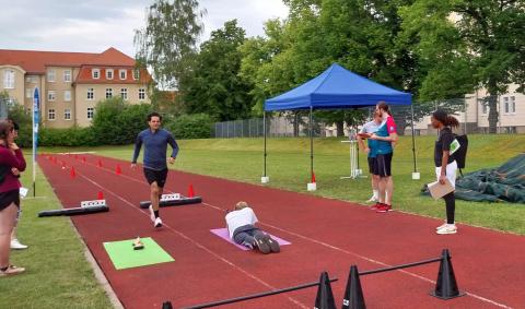 Ein Läufer auf einer Tartanbahn, auf der Nachbarbahn liegt eine Sportlerin mit einem Luftgewehr