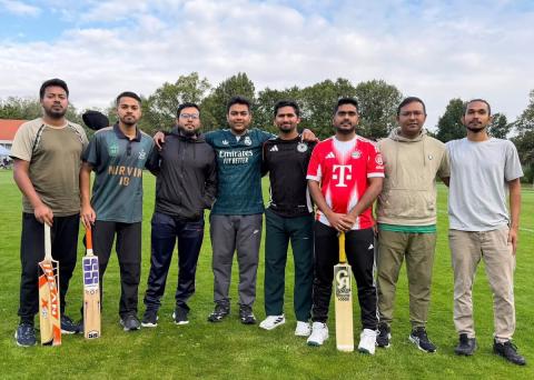 Gruppenbild einer Cricketmannschaft: 8 Studenten aus Bangladesch, drei davon mit Cricketschlägern auf einem Rasen-Sportplatz
