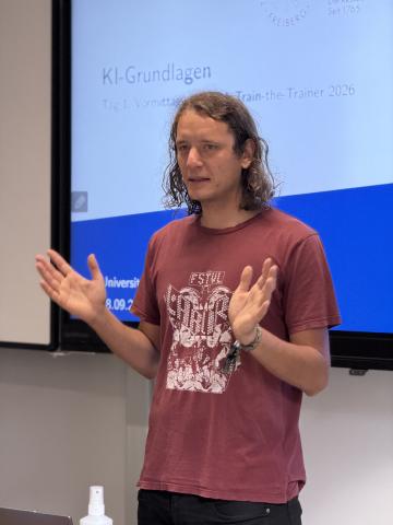 Mann mit schulterlangen Haaren und rotem T-Shirt gestikuliert vor einer Präsentationsfolie mit dem Titel 'KI-Grundlagen'