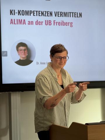Frau mit kurz geschnittenem Haar und Brille steht vor einer Präsentationsfolie und zeigt mit beiden Händen nach vorne