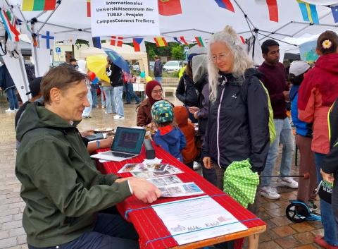An Mann sitzt an einem Stand mit zahlreichen Bildern darauf. Eine Besucherin des Standes betrachtet die Bilder. Im Hintergrund sieht man weitere Besucher und Standbetreuer.