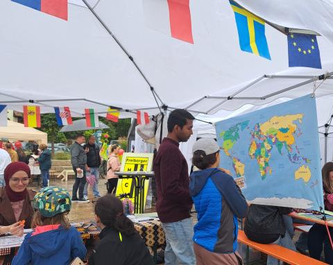 Ein internationaler Student steht mit einem Jungen vor einer farbenfrohen Weltkarte. Im Vordergrund ragen Länderfahnen als Wimpelkette vom Dach eines Pavillons.