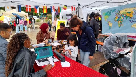 Eine Mutter mit Kind am einem einem Stand mit internationalen Studierenden. Im Hintergrund hängt eine farbenfrohe Weltkarte und Länderfahnen als Wimpelkette.