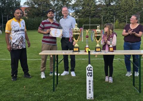 Fünf Personen stehen hinter einem Tisch auf dem sich 4 Sportpokale befinden. Ein weißer Cricketschläger lehnt vorn am Tisch.