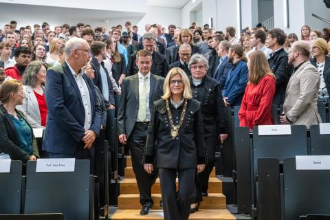 Festlich gekleidete Personen marschieren in einen gefüllten Hörsaal ein