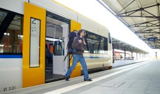 Ein überdachter Bahnsteig, ein Mann mit einem großen Rucksack steigt aus einem Zug mit gelben Türen.
