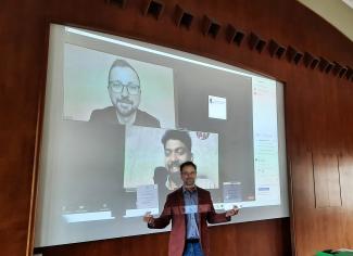 Dekan der Fakultät 4 der TUBAf zeigt vor einer Leinwand die Urkunden der beiden Preisträger, die online zugeschaltet sind. Die Gesichter beider Teilnehmer sind im Hintergrund auf der Leinwand zu sehen.