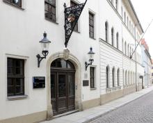 Ein helles, mehrstöckiges Gebäude (Altbau) mit eisernem Fahnenmast über dem bogenförmig geschwungenen Eingang mit der Aufschrift "Bergakademie" und den Symbolen des Bergbaus, Hammer und Eisen gekreuzt, darunter 2 altertümliche Straßenlaternen