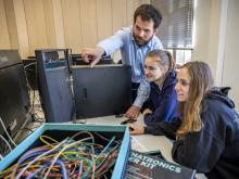 Zwei Studentinnen schauen auf einen Computer und ein Dozent weist auf etwas hin