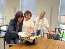 Drei Frauen stehen an einem Tisch und schauen sich ein altes Buch an.