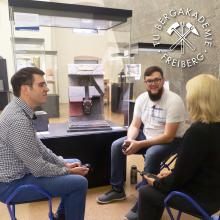 Drei sitzende Personen unterhalten sich in einem Ausstellungsraum.