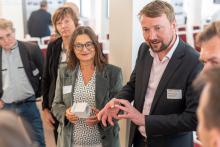 Festgesellschaft in einem Saal. Ein Mann erklärt Zuhörenden einen Gegenstand.