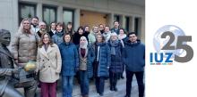 Ein Gruppenbild mit zahlreichen Männer und Frauen in Winterkleidung vor einem modernen Gebäude neben einer Statue von Alexander von Humboldt. Rechts neben dem Foto ein Logo mit einer Erdkugel und den Buchstaben IUZ sowie der Zahl 25.