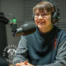 Eine junge Frau sitzt mit Kopfhörern vor einem Mikrofon und lächelt.