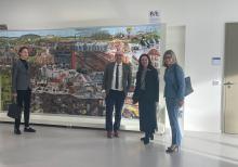 Eine Delegation aus vier Personen steht vor einem großformatigen Wimmelbild.