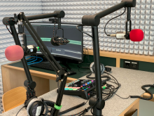 Podcaststudio mit zwei Mikrofonen an verstellbaren Armen, einem Mischpult mit bunten Tasten und einem Monitor auf einem Tisch.