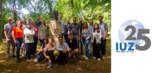 Ein Gruppenbild mit zahlreichen Männer und Frauen, die sich bei Sonnenschein in einem schattigen Park um eine Gedenksäule mit der Büste Alexander von Humboldts angeordnet haben. Rechts neben dem Foto zeigt ein Logo die Buchstaben IUZ und die Zahl 25 sowie einen stilisierten Globus (Vektorgrafik).